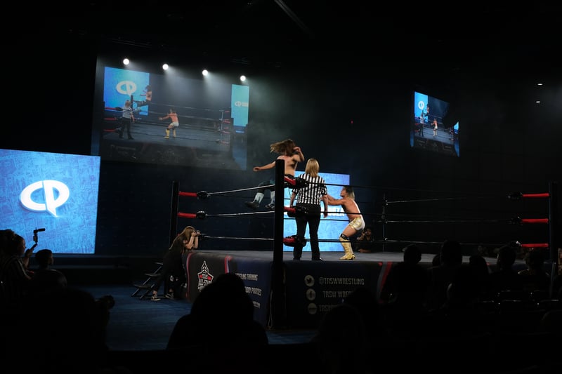 A wrestler falling backwards onto another as a referee looks on in the ring.