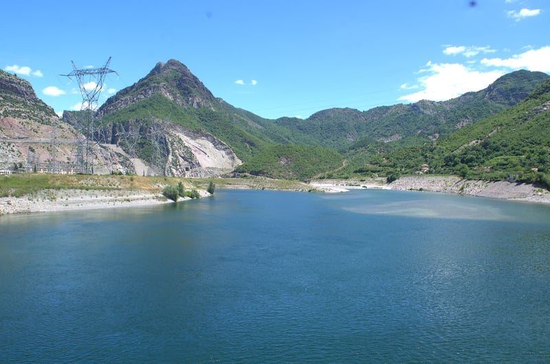 A blue Lake Koman in Albania with green hills and blue skies surrounding it.