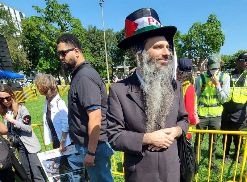 A man with a beard, long coat and Palestinian flag hat poses for a photo