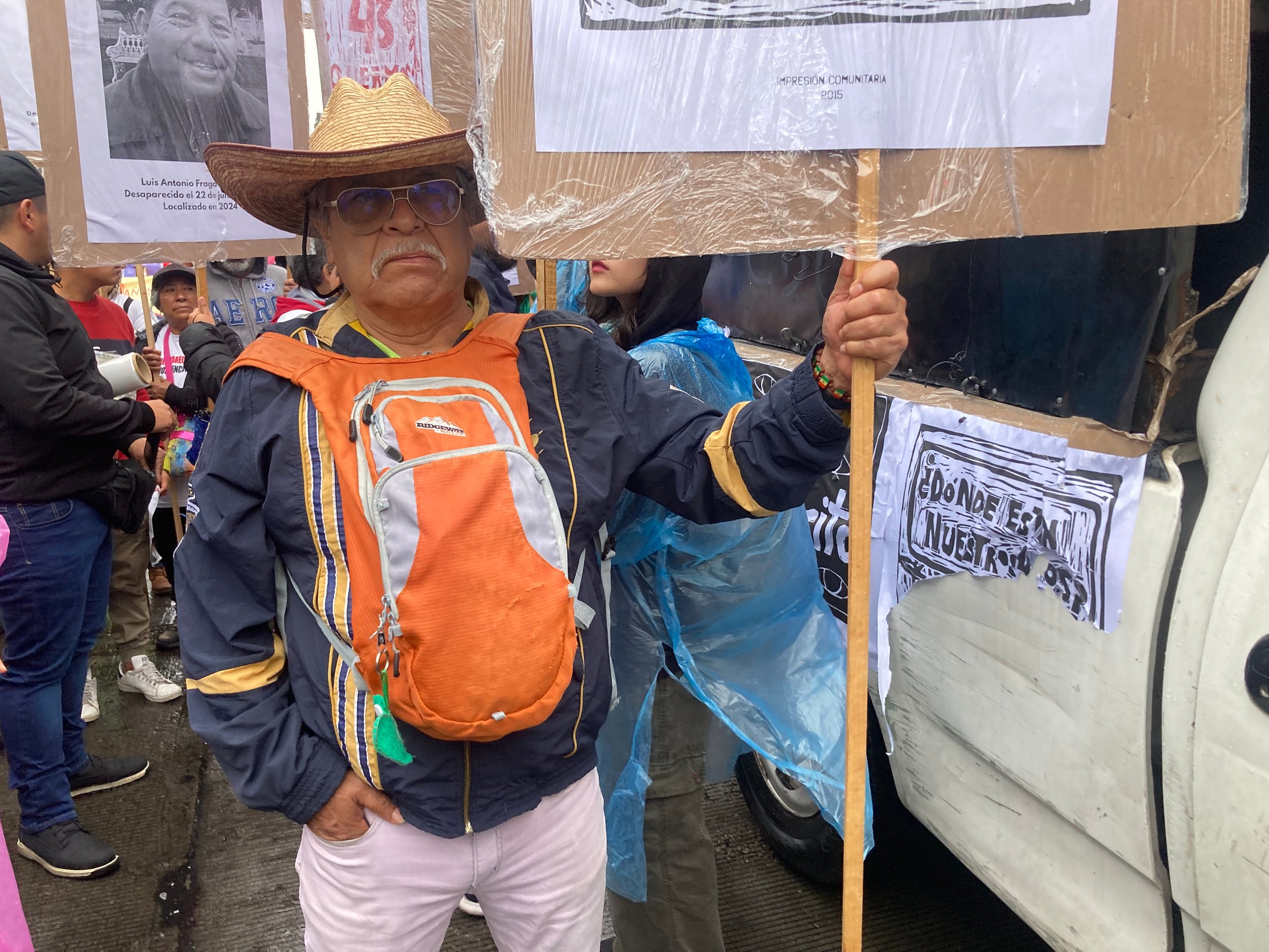 A man in a straw hat and an orange backpack on his chest holds a wooden stick attached to a sign in Mexico City.