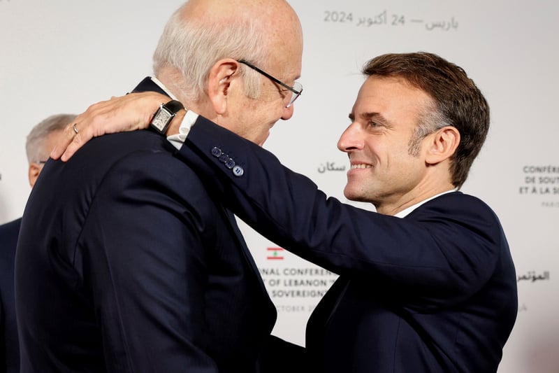 Emmanuel Macron hugs Najib Mikati in front of a background with French and Arabic words. Both men wear dark suits and Macron has a wristwatch visible as he lays his hand on Mikati's shoulder.