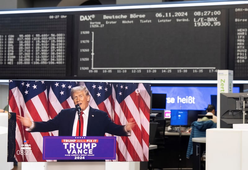 A TV showing Donald Trump with arms spread wide and a Trump/Vance placard on a podium, with the Deutsche Borse DAX signs behind.