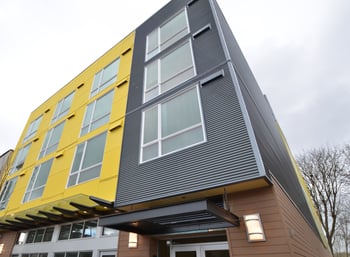 Modern apartment building exterior with bright yellow and grey corrugated siding and a covered en...