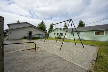 A playground with a swing set and a slide in the foreground and a green house in the background.
