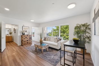 Living room with a white sofa, coffee table, and large window overlooking trees. Hallway to bathr...