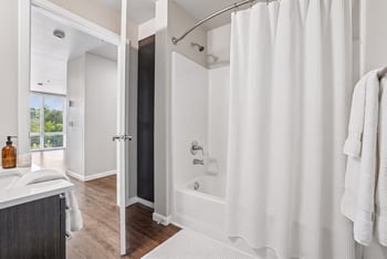Modern bathroom with white tub, curved shower rod, vanity counter, and view into the living space.