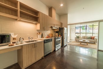 Modern kitchen with wood cabinetry and stainless appliances opening to a staged living area with polished concrete floors.