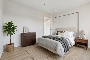 Staged bedroom with a bed, nightstand, dresser, potted plant, and sliding closet doors over light flooring.