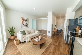 Staged living area with cream sofa, round coffee table, desk nook, and sliding doors leading to a bedroom alcove.