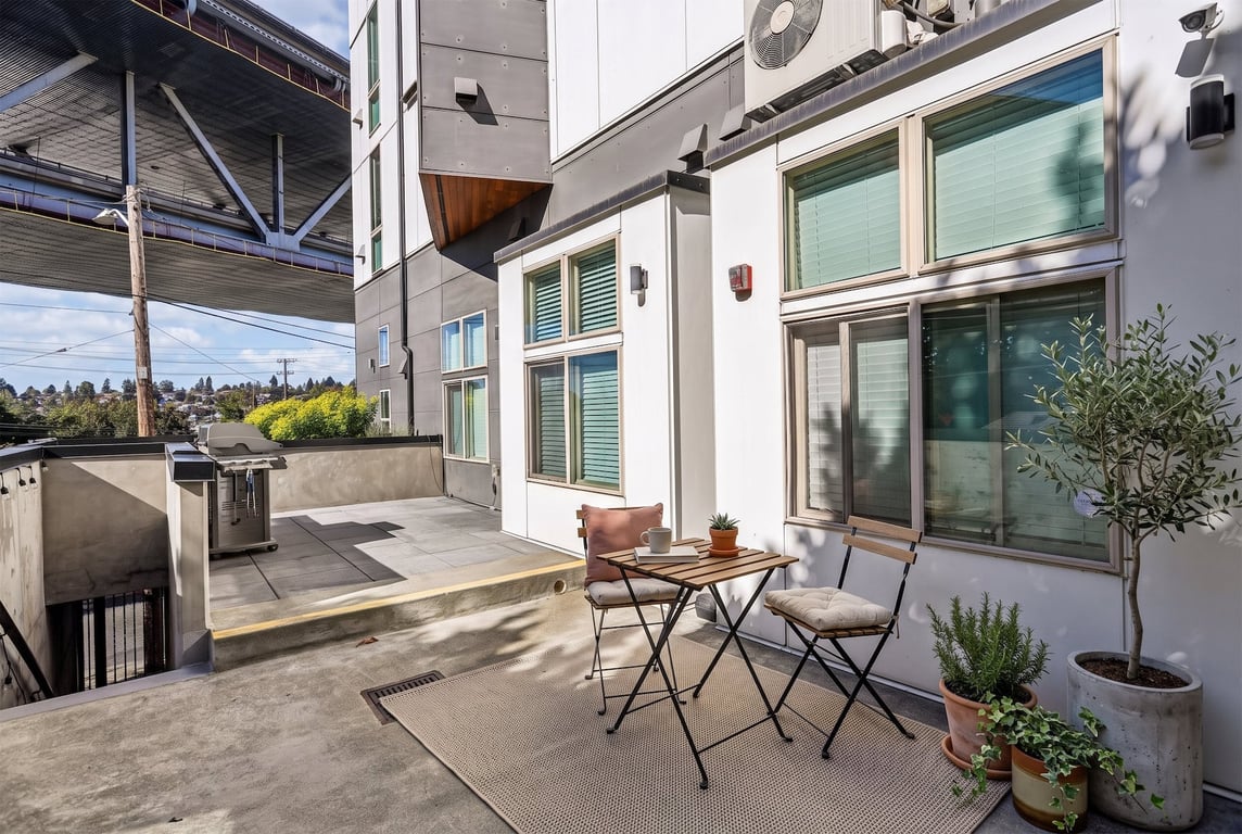 Outdoor patio with seating area, plants, and grill, next to an apartment building exterior with a...