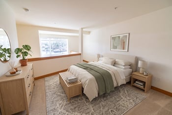 Loft bedroom with a made bed, dresser, mirror, and half-wall overlooking a bright window.