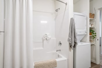 Bathroom with a white tub and shower and a stacked washer and dryer with built-in shelving alongside.