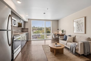 Open living area with sofa, coffee table, desk, and kitchen counter along the wall with a large window in the background.
