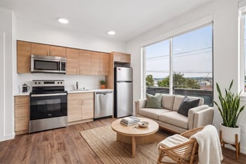 Open-concept living room and kitchen with wood-tone cabinets, stainless steel appliances, and a large picture window.