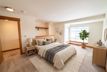 Bright carpeted bedroom with a double bed, wooden nightstands, dresser, and a large sunny window.