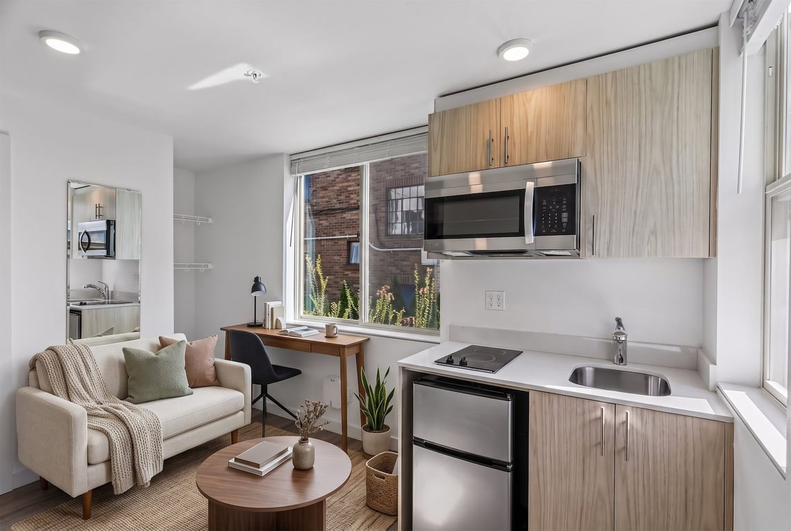 Studio apartment living space with a beige sofa, wood desk, and compact kitchen with stainless st...