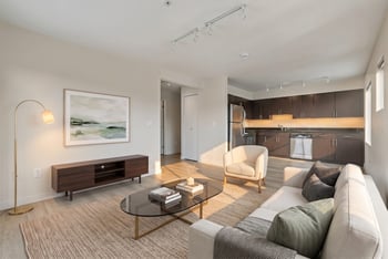 Staged living room with sofa, coffee table, and an open view into the kitchen with dark cabinets.