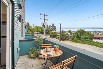 Private balcony with a round bistro table, two chairs, and potted herbs beside teal siding.