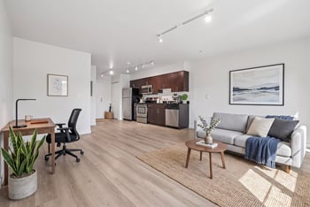 Open living area with wood-style flooring, sofa, desk, and kitchen with dark cabinets at REV Fremont.