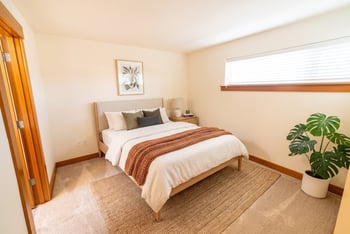 Bedroom with carpet, wood trim, bed with neutral linens, potted plant, and horizontal window with blinds.