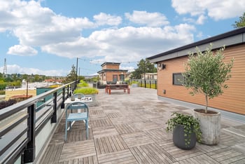 Rooftop deck with cafe chairs, a potted olive tree, and string lights strung along the railing.