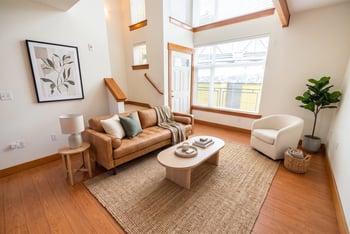 Staged loft living room with leather sofa, coffee table, white armchair, hardwood flooring, and tall double-height windows.