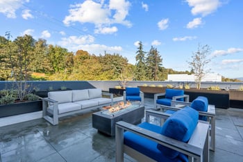 Rooftop patio with modern light sofa, blue chairs, a fire pit, and potted plants under a blue sky.
