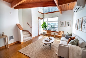 Loft living room with wood-beam ceiling, sofa, coffee table, stairs, and tall windows.