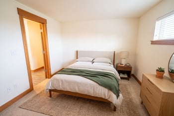 Staged bedroom with wood-frame bed, neutral bedding, green throw, nightstand, and dresser.