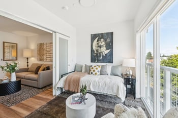 Living area with daybed, large window, and balcony access. Another room visible through sliding d...