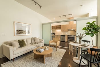 Staged living room with sofa, coffee table, small dining table, and sliding glass partition opening to the kitchen.