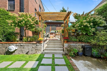 Outdoor stairs leading to a building entrance, covered by a wooden pergola, surrounded by lush ga...