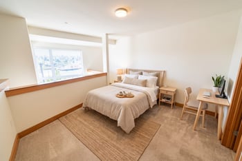 Loft bedroom with carpet, a made bed, wooden nightstand, desk with monitor, and a half-wall overlooking the lower level.