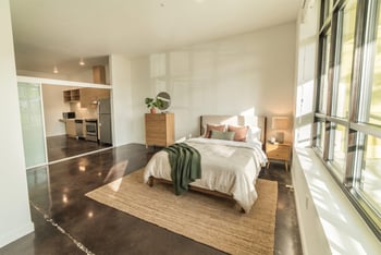 Staged studio bedroom with bed, dresser, concrete floors, large windows, and open view into the kitchen.