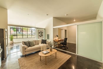 Staged living room with sofa, coffee table, polished concrete floors, and sliding partition to a work area.