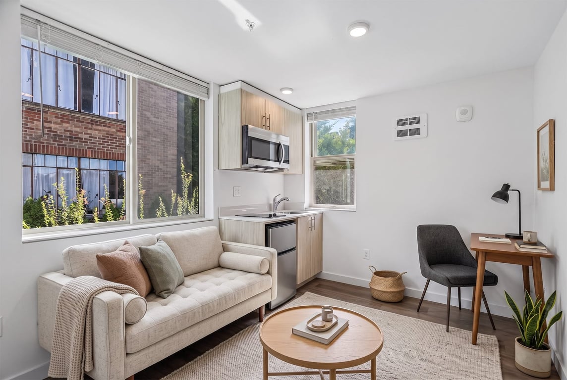Studio apartment with a sofa, desk, and compact kitchen featuring a microwave and mini-fridge.