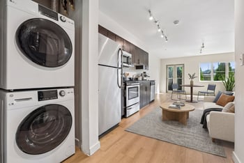 Open living area with stacked washer-dryer, linear kitchen with stainless appliances, and furnished sitting space.