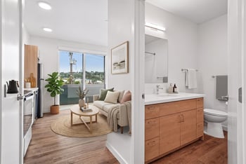 Apartment interior showing the kitchen, the living room with a view window, and a bathroom vanity with a wide mirror.