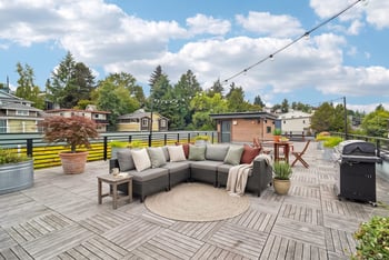 Rooftop deck with a sectional sofa, round rug, potted maple tree, and a gas grill under string lights.