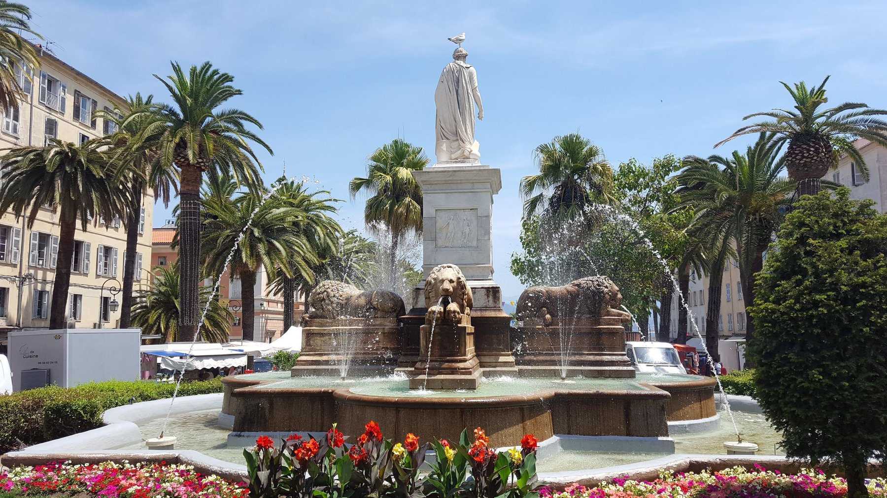 A water fountain surrounded by palm trees. A statue of a man sits in the centre, with a sea gull on its head.