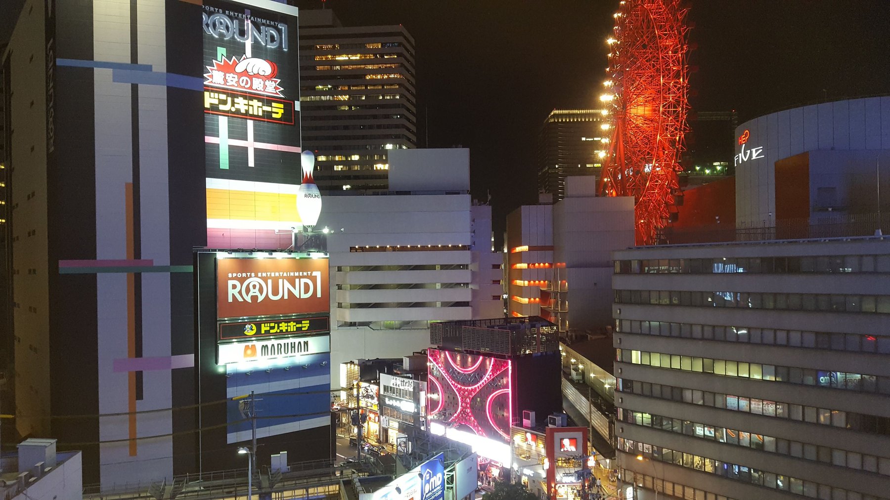 A large red ferris wheel, surrounded by tall buildings, lit up at night.