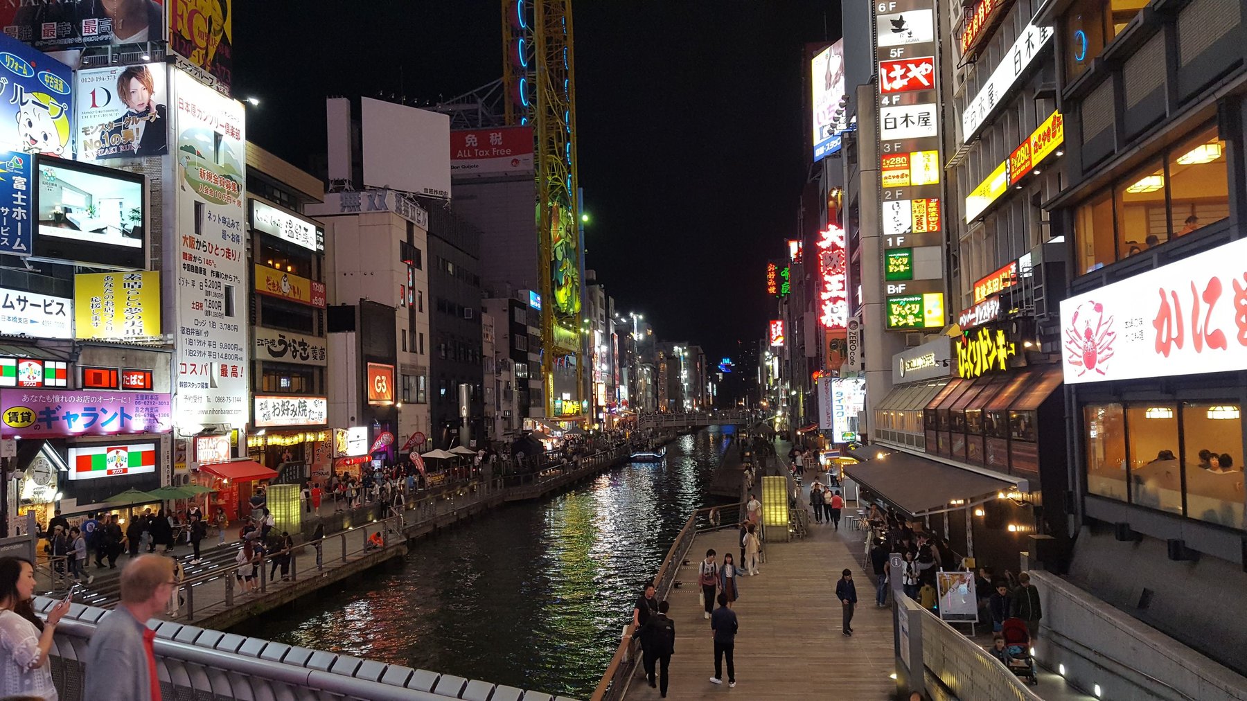 Shops and restaurants with illuminated signs line boardwalks either sides of a narrow river.