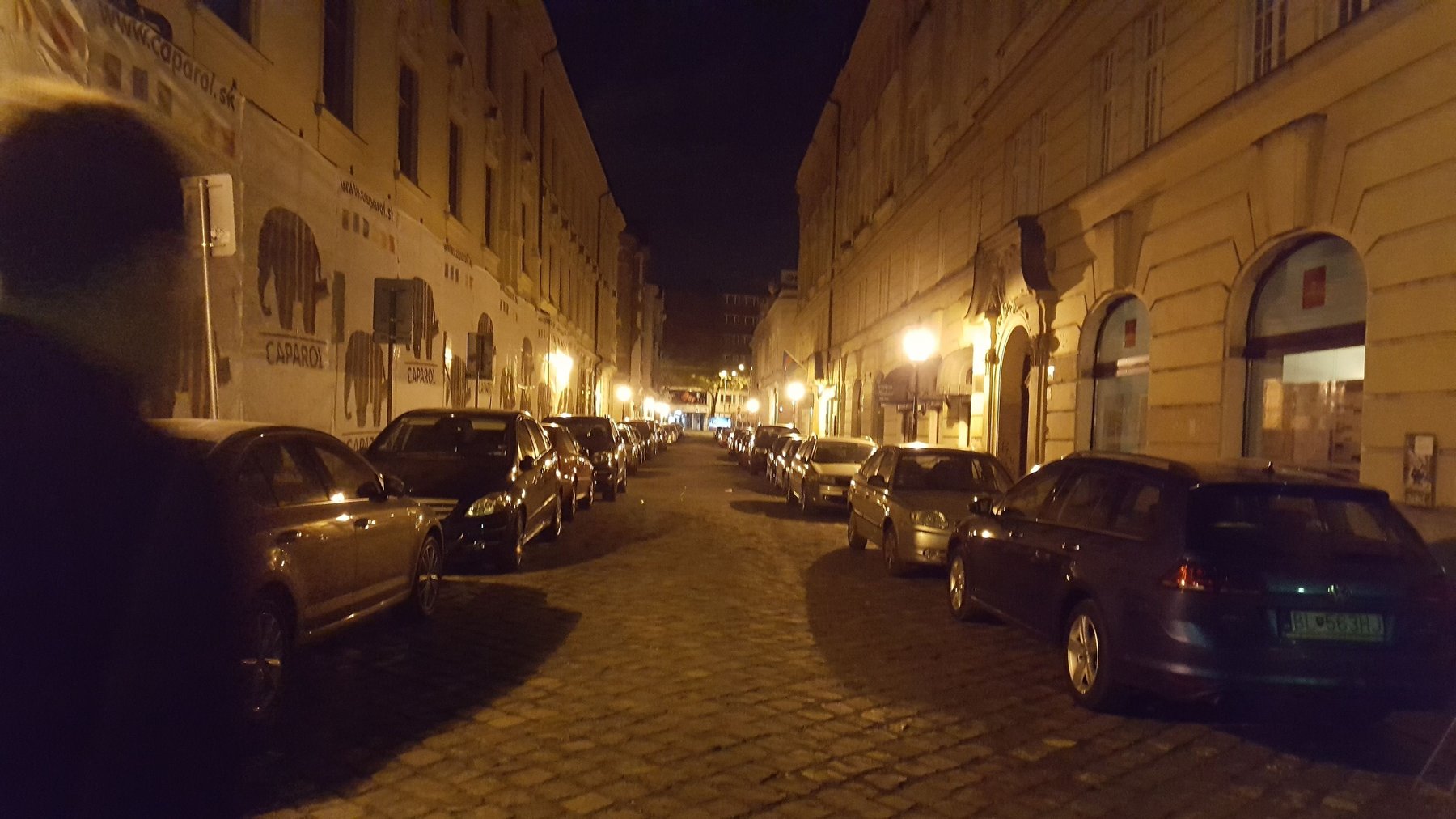 A cobbled street at night, lined with cars.