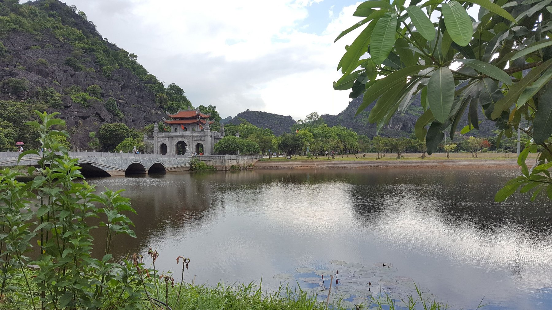 A bridge leading to an ornate gate viewed from across a still river.