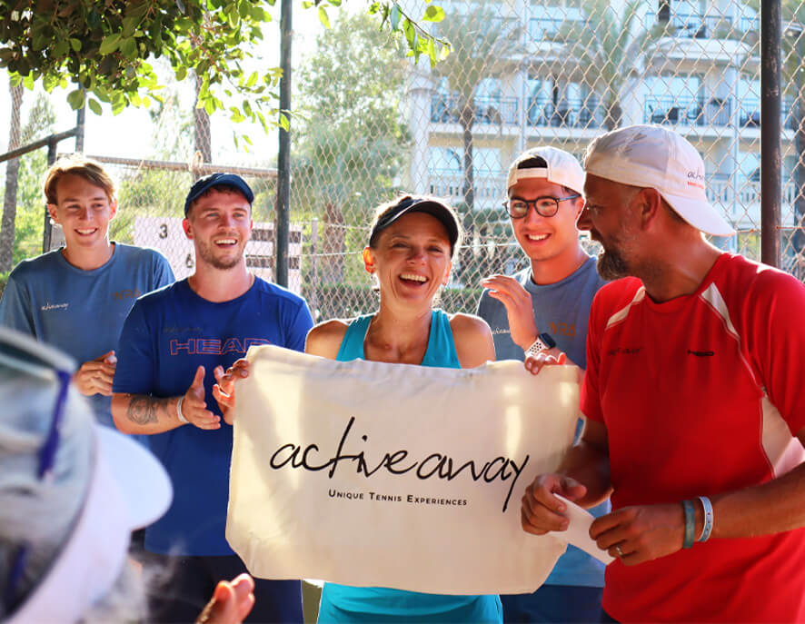 Group of adults smiling on a tennis court, holding an 'active away' banner during a hosted tennis holiday experience.