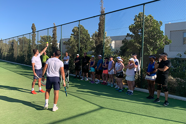 Tennis coach demonstrating a technique to players on an outdoor court with a group of participants watching closely.