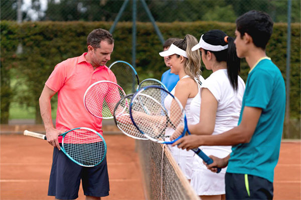 Tennis coach guiding three students at the net on a clay court, each holding a tennis racket.