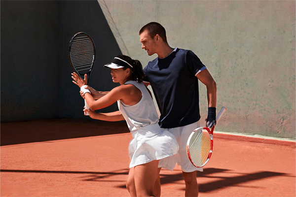 A man and woman playing doubles tennis on a clay court, the woman preparing a backhand stroke with her partner observing.