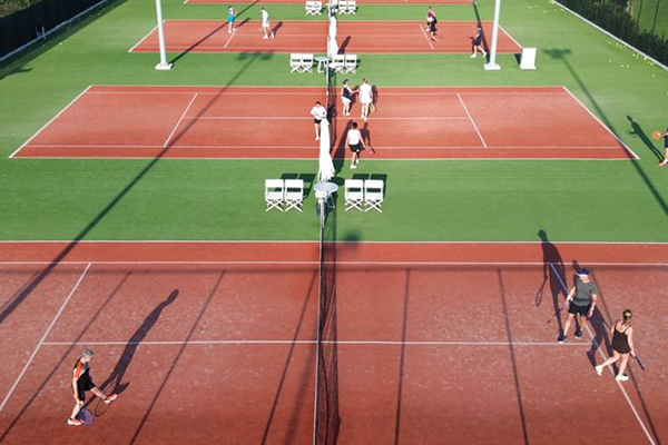 Overhead view of multiple clay tennis courts with players practicing and playing matches, casting long shadows in the late afternoon light.