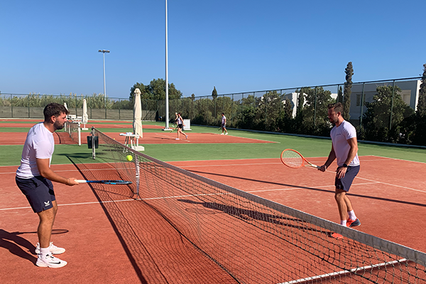 Two male tennis players practising on a red clay tennis court with clear blue skies, surrounded by trees and a fence, while other players are in the background.